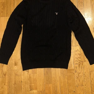 Svart kabelstickad tröja från GANT - Säljer en stilren svart kabelstickad tröja från GANT i premium bomull. Tröjan har långa ärmar och ett diskret broderat GANT-emblem på bröstet. Perfekt för en klassisk och bekväm look.   Passar även som L