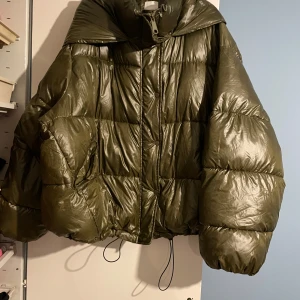 Olivgrön pufferjacka från H&M - Säljer en olivgrön pufferjacka från H&M. Jackan har en glansig yta och är quiltad med dragkedja framtill. Perfekt för kyligare dagar med sin varma och puffiga design. Den har en hög krage och justerbar nederkant. Jackan har ett hål med lagningslapp, men det syns inte så mycket pga oversize modell. 