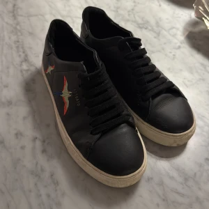 ARIGATO - Snygga svarta sneakers från Axel Arigato med ett färgglatt fågelbroderi på sidan. Skorna har en rund tå och klassisk snörning. Perfekta för en stilren look.