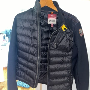 Svart jacka från Parajumpers - Snygg svart jacka från Parajumpers med quiltad design och dragkedja framtill. Jackan har en cool patch på armen och en gul detalj vid kragen. Perfekt för kyligare dagar. Jag valde att sälja denna underbara jacka eftersom att jag har vuxit ur den