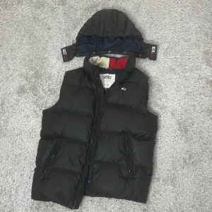 Svart dunväst från Tommy Hilfiger i storlek M - Snygg svart dunväst från Tommy Hilfiger med avtagbar huva. Västen har dragkedja och två fickor med dragkedjor. Insidan är fodrad i blått med röda och vita detaljer. Perfekt för kyligare dagar. Storlek M Passar till 170-180cm