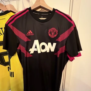 United  träningströja  - Snygg svart fotbollströja från Adidas med röda detaljer. Tröjan har korta ärmar och är tillverkad med Climalite-material för optimal komfort. Perfekt för fotbollsfans som vill ha en stilren look.