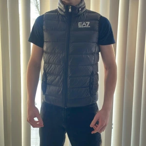 Svart dunväst från Emporio Armani - Säljer en stilren svart dunväst från Emporio Armani EA7. Västen har en dragkedja framtill och två praktiska fickor. Perfekt för att hålla värmen med stil. Nypris 1399kr, Passar bra till både casual och mer uppklädda tillfällen. Bra pris!!✅