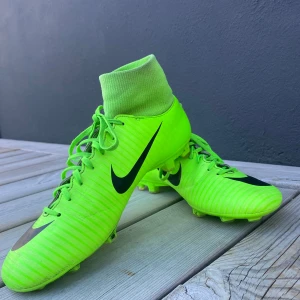 Neongröna fotbollsskor från Nike - Säljer ett par neongröna Nike fotbollsskor med hög ankelstrumpa och snörning. Skorna har en svart Nike-logga på sidan och är designade för att ge bra grepp på planen. Perfekta för fotbollsspelare som vill sticka ut på planen.