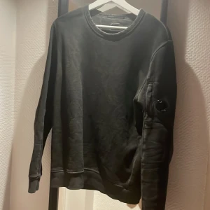 Svart sweatshirt från C.P. Company - Säljer en stilren svart sweatshirt från C.P. Company. Tröjan har en rund halsringning och en unik detalj med en lins på ärmen. Perfekt för en casual look. Jag säljer den för att den har vuxit ur mig, tyvärr. Ny pris: 2000kr. Köpt på Cenino i Växjö!