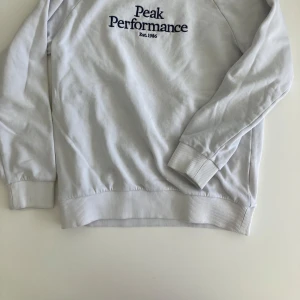 Vit tröja från Peak Performance - Säljer en vit tröja från Peak Performance med broderad logga på framsidan. Tröjan har långa ärmar och ribbade muddar vid ärmslut och nederkant. Perfekt för en avslappnad stil.