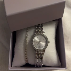 Silver klocka & armband - Elegant silverfärgad klocka från regal med glittrande stenar runt urtavlan. Klockan har en länkarmband och en stilren design som passar perfekt för festliga tillfällen. Urtavlan är i en ljus silverton med tydliga markeringar.