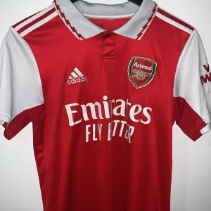 Röd Arsenal fotbollströja från Adidas - Säljer en röd Arsenal fotbollströja från Adidas med vit krage och korta ärmar. Tröjan har klubbens emblem på bröstet och sponsorlogga. Namnet 'Zinchenko' och nummer 35 är tryckt på ryggen. Perfekt för alla Arsenal-fans! ⚽️