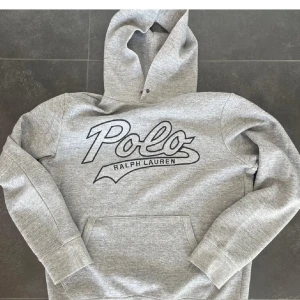 Grå hoodie från Ralph Lauren - Säljer en stilren grå hoodie från Ralph Lauren med stor logga på bröstet. Tröjan har en klassisk känguruficka och en bekväm huva med dragsko. Perfekt för en avslappnad look. Liten i storlek 