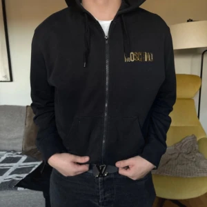 Svart hoodie från Moschino - Säljer min killes äkta kofta från Moschino med dragkedja och guldfärgad logga på bröstet. Köptes för 5000 och nästan helt oanvänd 