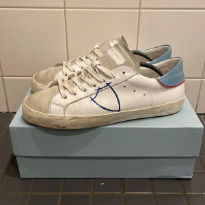 Philippe Model - Snygga vita sneakers från Philippe Model med blå och röda detaljer. Skorna har en rund tå och snörning framtill. De är tillverkade i skinn med en gummisula och har en stilren design med en blå detalj på sidan. Skorna är 100% Autentiska och pris kan diskuteras! BOX INGÅR!