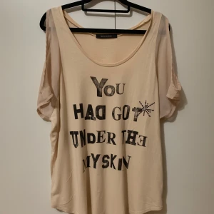 Beige t-shirt med tryck från Vence Exchange  - Säljer en beige topp med texten 'You Had Got Under My Skin' i svart och glittriga detaljer. Toppen har korta ärmar och en lös passform. Perfekt för en avslappnad stil.