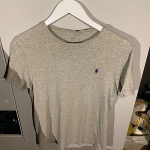 Grå t-shirt från Ralph Lauren - Säljer en stilren grå t-shirt från Ralph Lauren med en liten blå logga på bröstet. T-shirten är kortärmad och gjord i mjuk bomull, perfekt för en avslappnad look. Passar bra till jeans eller shorts! Passar 170-175 bäst!