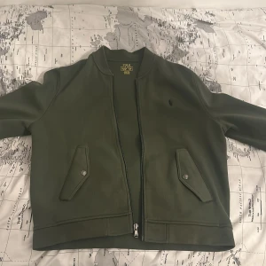 Grön bomberjacka från Ralph Lauren - Säljer en stilren grön bomberjacka från Ralph Lauren. Jackan har en dragkedja framtill och två fickor med knappar. Perfekt för en avslappnad look.