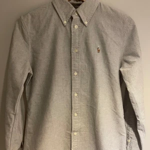 Grå skjorta från Ralph Lauren - Säljer en stilren grå skjorta från Polo Ralph Lauren med broderad logotyp på bröstet. Skjortan har en lite defekt på höger Axel som syns på bild! .Perfekt för sommaren! pris går självklart att diskuteras.