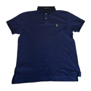 Ralph Lauren piké  - Säljer en klassisk mörkblå pikétröja från Ralph Lauren med gul broderad logga på bröstet. Tröjan har korta ärmar och knappar vid kragen. Perfekt för en stilren look! Få mörka fläckar på bröstet, inte så tydliga. Storlek L 