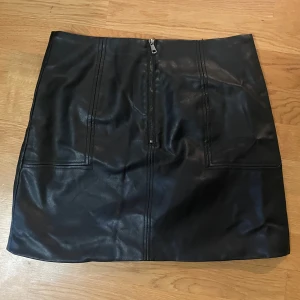 Svart skinnkjol med dragkedja - Snygg svart skinnkjol med dragkedja framtill. Perfekt för en cool och stilren look. Kjolen har en kort längd och är gjord i ett mjukt skinnmaterial.