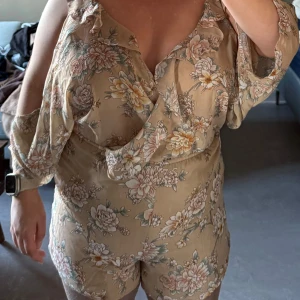 Ljusblommig byxdress från Forever 21 med v-ringning, volangdetaljer och öppen axel. Superfin till sommar, fest, semester eller midsommarfirande! -  Storlek: M (passar även L tack vare stretch i midjan) Skick: Väldigt fint skick – använd 1 gång. Färg: Beige med blommor i rosa, vitt och grönt Detaljer: 	•	Lätt tyg som faller fint 	•	Resår i midjan 	•	3/4-ärm med “cold shoulder”-öppning 	•	Kort modell (shorts)  Passar perfekt med sandaler, sneakers eller klackar