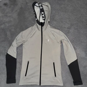 Grå huvtröja från Peak Performance - Snygg grå huvtröja från Peak Performance med svarta detaljer och dragkedja. Tröjan har långa ärmar och en justerbar huva med logotyp. Perfekt för en sportig look eller avslappnade dagar. Pris kan diskuteras 