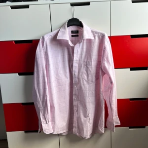 Rosa rutig skjorta från Thomas Nash - Säljer en stilren rosa skjorta från Thomas Nash i 100% bomull. Skjortan har ett rutigt mönster med blå linjer och en bröstficka. Perfekt för en klassisk look.