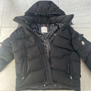 Svart dunjacka från Moncler - Säljer en snygg svart dunjacka från Moncler med huva och dragkedja. Jackan har en praktisk ficka på ärmen med knappstängning och Moncler-logga. Perfekt för kalla dagar!