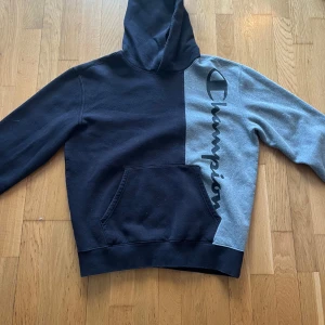 Svart och grå hoodie från Champion - Säljer en cool hoodie från Champion i svart och grått med stor logga på framsidan. Tröjan har en klassisk känguruficka och huva. Perfekt för en avslappnad stil.