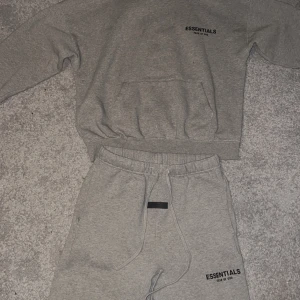 Grå hoodie från Essentials - Säljer en grå hoodie från Essentials med tryck på bröstet. Tröjan har en klassisk känguruficka och ribbade muddar vid ärmslut och nederkant. Perfekt för en avslappnad stil.