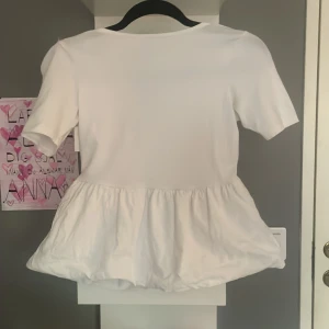 Vit blus med volangdetalj - Säljer en söt vit blus med korta ärmar och volangdetalj i nederkant. Perfekt för en somrig look. Blusen har en rund halsringning och är gjord i ett mjukt material.