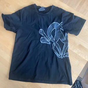 Svart t-shirt med tryck från Syna - Säljer en svart t-shirt från Syna World med ett unikt grått tryck på framsidan. T-shirten har en normal passform och är kortärmad. Perfekt för en avslappnad stil. vid snabb affär kan priset sänkas. vid frågor hör av dig