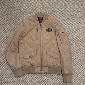 Beige quiltad bomberjacka från Morris - Snygg beige quiltad bomberjacka från Morris med ribbade muddar och dragkedja. Jackan har en broderad detalj på bröstet och en ficka med dragkedja på ärmen. Tveka inte över att höra av er vid frågor eller funderingar!☺️
