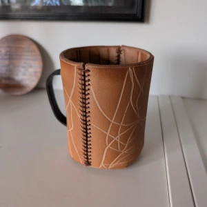 Handgjord mugg med läderomslag - Unik handgjord mugg med ett snyggt läderomslag i brunt med vita linjemönster. Muggen har en svart handtag och dekorativa sömmar längs sidan. Perfekt för att ge en rustik känsla till din kaffestund.