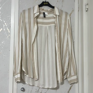 Vit och beige randig skjorta - Snygg vit skjorta med beige ränder från H&M. Skjortan har långa ärmar och knappar framtill. Perfekt för en stilren look.