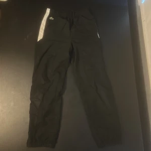 Svarta träningsbyxor från Lacoste - Snygga svarta träningsbyxor från Lacoste med vita detaljer längs sidorna och elastisk midja. Byxorna har det klassiska Lacoste-logotypen på framsidan och är perfekta för sportiga aktiviteter.  Texten på sidan är aningen sliten, se bild 5.