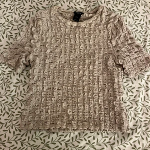 Beige topp från Lindex - Säljer en unik beige topp från Lindex med ett texturerat, rynkat mönster. Toppen har korta ärmar och en rund halsringning. Perfekt för att ge din outfit en intressant touch. 🌟