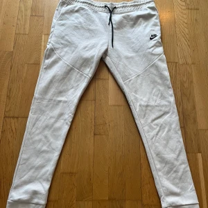 Beiga mjukisbyxor från Nike - Säljer ett par vita mjukisbyxor från Nike med svart snörning i midjan och en diskret logga på låret. Byxorna har en slim passform och är perfekta för en avslappnad stil. De har även en praktisk ficka med dragkedja på sidan.