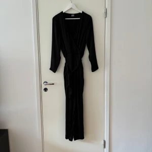 Svart långklänning från Gina Tricot - En elegant svart jumpsuit från Gina Tricot. Jumpsuiten har långa ärmar och en omlott-design framtill. Den är tillverkad i ett mjukt och följsamt material som ger en fin passform.