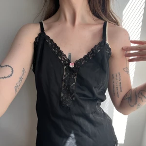 Svart satinlinne med spets och ros, vintage - Supergulligt svart linne, med en liten ros i fram och fina spetsdetaljer. Vintage och i fint skick! Jag själv har aldrig använt det. Storlek large enligt storlekstag, men passar mer en S!