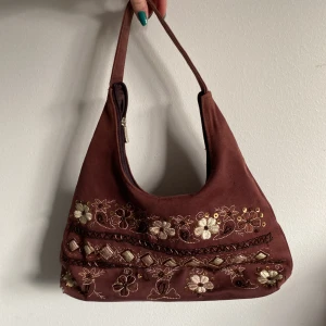 Brun handväska med paljetter och pärlor - Broderad handväska med blommönster, pärlor och paljetter. Blixtlås och ett innerfack. Bra skick med lite slitage på bottnen samt ett par lösa trådar och pärlor, kan skicka mer bilder vid önskemål!