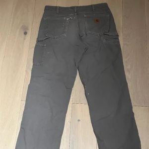 Grå Carthartt carpenter pants - Säljes gråa carhattbyxor. De har lite färgstänk enligt bilder och är klippta från att vara 36 tum långa till 34. Fler bilder kan absolut fixas!