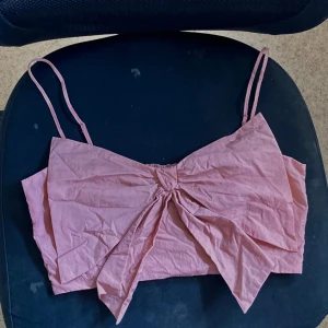 Topp med en rosett 🎀 - En kort topp i taft med stor, dekorativ rosett fram. Toppen har smala, reglerbara axelband och smockat parti bak. Fodrat framstycke.🎀🫧✨  Längd: Kort Ärmlängd: Ärmlös Modell: Smock, Korsettopp Beskrivning: Puderrosa, Enfärga