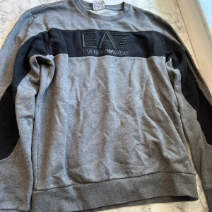 Grå/svart Emporio Armani tröja  - En grå/svart emporio Armani tröja i storlek XS/S. Tröjan sitter relativt tajt och har ett mjukt material. Tröjan har ett stort EA7 tryck i mitten men svarta detaljer på ärmarna. Tröjan har använts ett par gånger under året men ser samma ut.