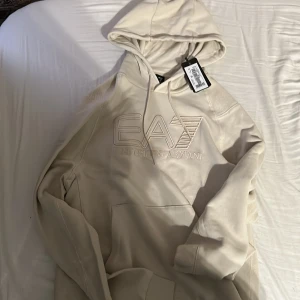 EA7 Beige Hoodie - Denna EA7 hoodie är i nyskick, med prislappen fortfarande på. Nypriset på denna är cirka 1500kr och jag säljer den nu då jag inte gillade den. Storleken är M