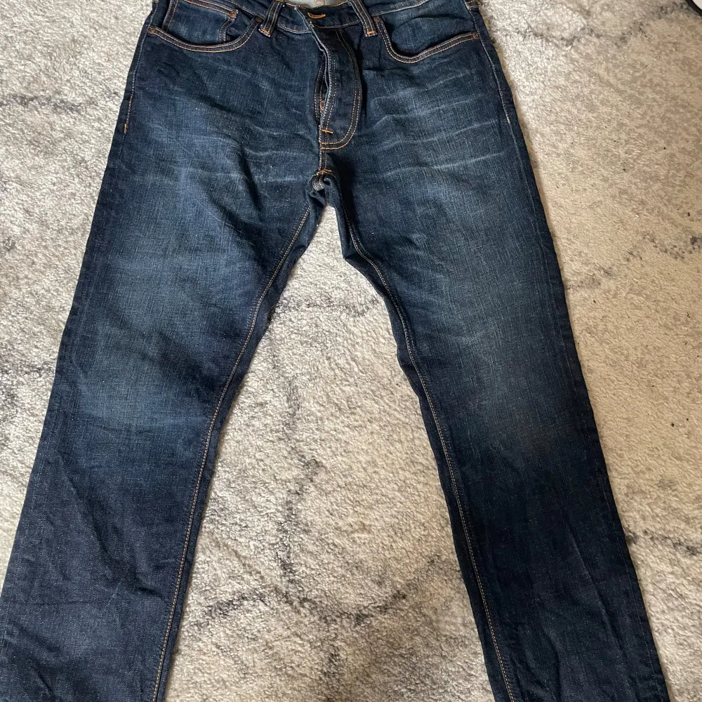 Ett par mörkblå jeans från Nudie Jeans med modellen Lean Dean Troubled Sea. Jeansen har en knappgylf och orange sömmar. Passar storlekarna M/L.. Jeans & Byxor.