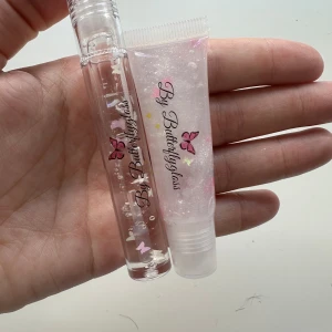 By Butterflygloss Läppglans - Två läppglans från By Butterflygloss. Ett i en genomskinlig tub med skruvlock och ett i en genomskinlig klämtub. Båda har en genomskinlig bas med glitter och fjärilsdekorationer på förpackningen. Priset gäller båda läppglansen! 