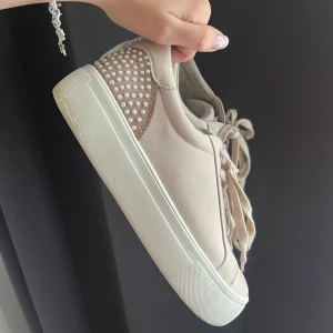 Beige sneakers från Anna Field - Ett par beige sneakers från Anna Field med snörning och dekorativa pärlor på hälen. Skorna har en vit sula och är tillverkade i syntetmaterial.