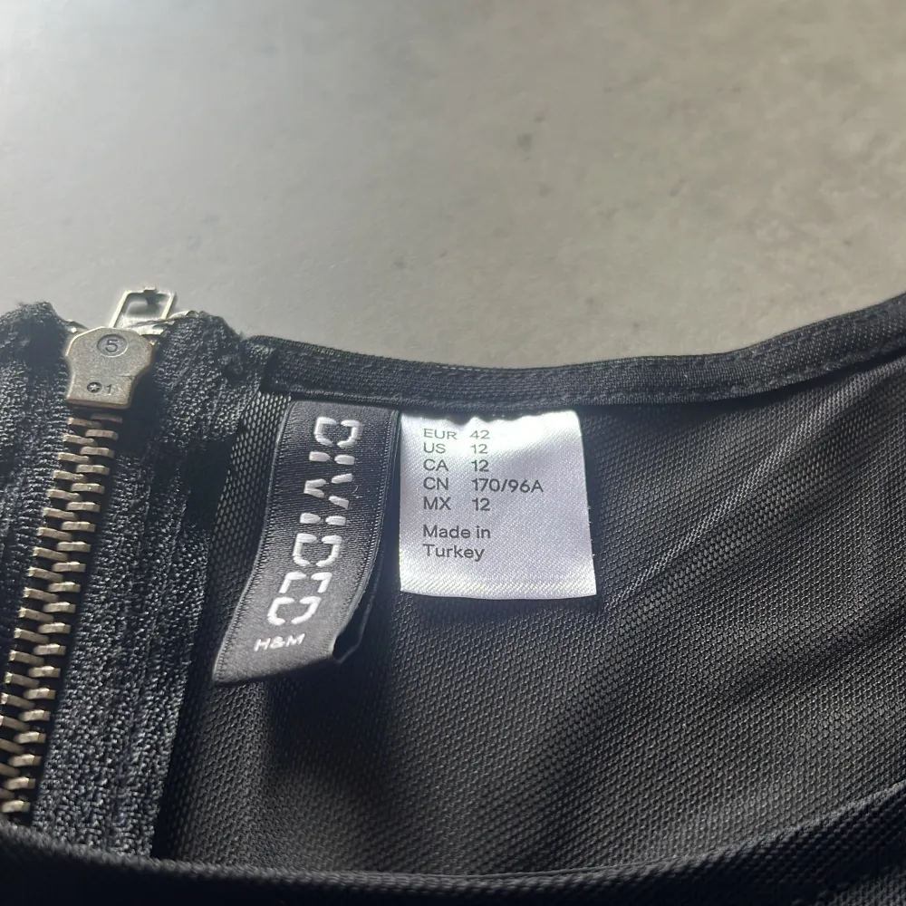 En elegant svart klänning från H&M Divided med trekvartsärm och dragkedja i ryggen. Klänningen har en transparent mesh-detalj på ärmarna och övre delen av ryggen.. Mekot.