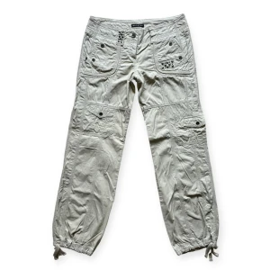 Vintage Watcher lågmidjade beige cargobyxor - Beige cargo byxor med dragkedja och knapp. Byxorna har flera fickor och detaljer i form av nitar. Byxorna har en smal passform och är tillverkade i ett lätt och bekvämt material. Helt oanvända, köpta 2010 i Amsterdam. 