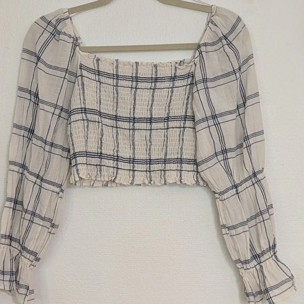 En rutig blus med långa ärmar. Blusen har en elastisk, rynkad design på kroppen med ihopdragningar på ärmarna. croptop. Färgerna är vit och blå rutigt. Toppar.