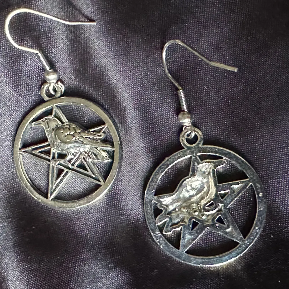 Ett par unika silverfärgade örhängen. Örhängena har en rund design med ett pentagram i mitten och en korp ovanpå på pentagrammet. 4,6 cm långa, 2,4 cm breda. Allergivänliga krokar, gummistoppar medföljer. Tillverkade av mig @bybasen. Uppseendeväckande och ovanliga. Unisex. Bundlerabatt.. Asusteet.