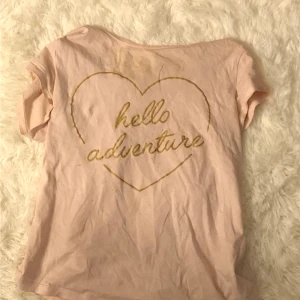 En ljus rosa tröja med tryck - En ljusrosa kortärmad med ett hjärta i guld och inne i hjärtat står de hello adventure 